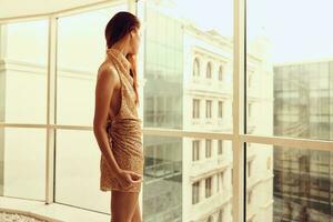 Woman in gold sequined dress standing by the window posing in a stylish look with long hair, designer dress with open back, party look photo