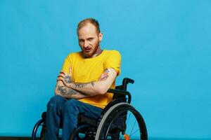 un hombre en un silla de ruedas en un camiseta con tatuajes en su brazos se sienta en un azul estudio fondo, un lleno vida, un real persona foto