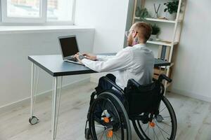 A man in a wheelchair looks at the camera businessman in the office working on a laptop online, social networks startup, integration into society, concept working person with disabilities, view back photo