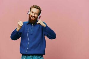 Portrait of a redheaded man wearing headphones smiling and dancing, listening to music on a pink background. A hipster with a beard. photo