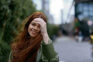 Portrait of a beautiful young woman in the city looking into the camera smile with teeth with red flying hair in fashionable clothes in the city against of bamboo in spring, lifestyle in the city photo