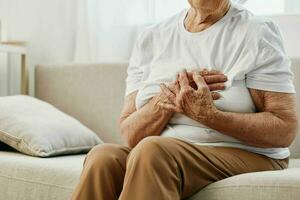 Elderly woman severe chest pain sitting on the sofa, health problems in old age, poor quality of life. Grandmother with gray hair holds her chest with her hands, women's health, breast cancer. photo