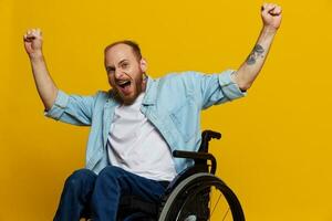 un hombre en un silla de ruedas sonrisa y felicidad, pulgar arriba, con tatuajes en su manos se sienta en un amarillo estudio fondo, el concepto de salud un persona con discapacidades foto