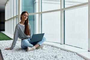 A woman works online via the Internet on her laptop and smiles looking out the large windows of a tall building, an urban style concept and working in an office photo