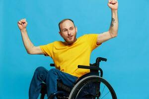 A man in a wheelchair happiness raised his hands up in a t-shirt with tattoos on his hands sits on a blue studio background, a full life, a real person photo