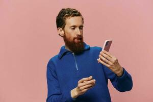 Portrait of a redheaded man with phone in hand taking selfies and photos on his phone with a smile on a pink background, blogger