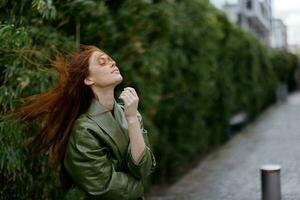 Fashion woman in the city on the street posing in front of buildings and trees with red long flyaway hair in the windy weather photo