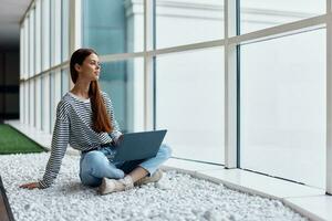 A woman works online via the Internet on her laptop and smiles looking out the large windows of a tall building, an urban style concept and working in an office photo