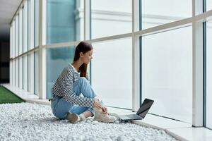 A woman smiling with teeth sits with a laptop by the big windows of a building in the city, the concept of work and leisure in the big city photo
