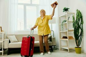 Happy senior woman with passport and travel ticket packed a red suitcase, vacation and health care. Smiling old woman joyfully stands in the house before the trip. photo
