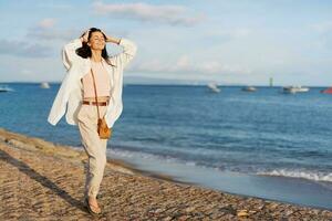 un mujer contento con su ojos cerrado en el Dom caminando en el playa con su brazos untado fuera en contra el Oceano con un sonrisa, volador cabello, bronceado piel, relajación, un viaje a el Oceano y libertad. foto