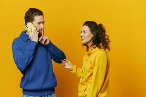 Man and woman couple with phone in hand call talking on the phone, on a yellow background, symbols signs and hand gestures, family quarrel jealousy and scandal. photo