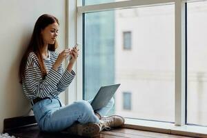 Happy woman sitting on the floor by the windows with her laptop and holding her phone looking at it, a view of the big city, the concept of working and learning online photo