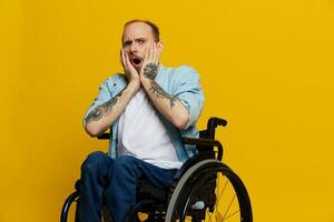 un hombre en un silla de ruedas sorprendido, con tatuajes en su brazos se sienta en un amarillo estudio fondo, el concepto de salud es un persona con discapacidades foto