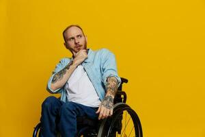 A man in a wheelchair problems with the musculoskeletal system looks at the camera thoughtfully with tattoos on his arms sits on a yellow studio background, health concept a person with disabilities photo