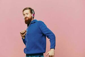 Portrait of a redheaded man wearing headphones smiling and dancing, listening to music on a pink background. A hipster with a beard. photo