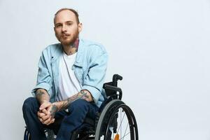 A man in a wheelchair looks thoughtfully at the camera, copy space, with tattoos on his arms sits on a gray studio background, the concept of health a person with disabilities, a real person photo