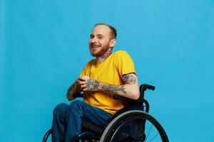 un hombre en un silla de ruedas sonrisa mira a el cámara en un camiseta con tatuajes en su brazos se sienta en un azul estudio fondo, un lleno vida, un real persona foto