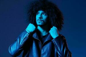 Portrait of fashion man with curly hair on blue background multinational, colored light, black leather jacket trend, modern concept. photo