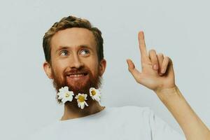 Portrait of a funny man in a white T-shirt with flowers daisies in his beard on a white isolated background, copy place. Holiday concept and congratulations. photo