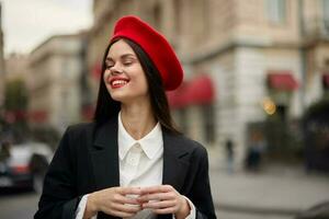 Moda mujer retrato sonrisa con dientes en pie en el calle en frente de el ciudad turista en elegante ropa con rojo labios y rojo boina, viajar, cinematográfico color, retro Clásico estilo, urbano moda. foto