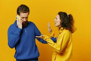 Man and woman couple with phone in hand call talking on the phone, on a yellow background, symbols signs and hand gestures, family quarrel jealousy and scandal. photo