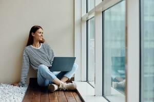 Business woman sitting with a laptop at the window full-length and smiling with teeth and looking out the window, happy woman work online in the office, copy place photo