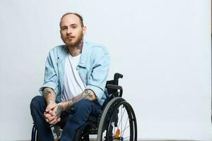 A man in a wheelchair looks thoughtfully at the camera, copy space, with tattoos on his arms sits on a gray studio background, the concept of health a person with disabilities, a real person photo