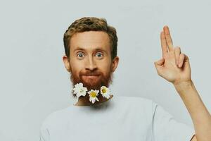 Portrait of a funny man in a white T-shirt with flowers daisies in his beard on a white isolated background, copy place. Holiday concept and congratulations. photo