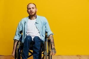 A man in a wheelchair looks at the camera, with tattoos on his arms sits on a yellow studio background, the concept of health is a person with disabilities photo