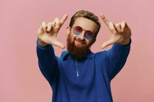 Portrait of a redheaded man wearing sunglasses smiling and dancing, listening to music on a pink background. Hipster with a beard, happiness finger pointing. photo