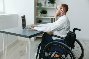 A man wheelchair businessman with tattoos office works at a laptop online, social networks and startup, integration into society, the concept of working a person with disabilities, view from the back photo