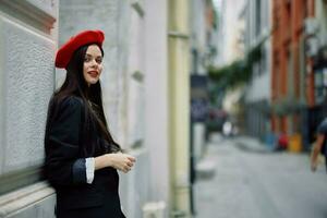 un mujer en pie cerca un pared en el ciudad vistiendo un elegante chaqueta y un rojo boina con rojo labios, de viaje. foto