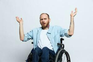 A man in a wheelchair and pointing a finger at, copy space, with tattoos on his hands sits on a gray studio background, health concept a person with disabilities, a real person photo