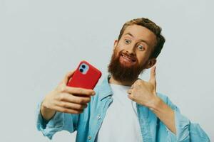 Man hipster with phone in hand blogger smile on gray background in blue shirt and white t-shirt, talking on the phone and selfies photo