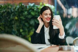 elegante Moda mujer blogger turista se sienta en un café con un teléfono en su manos lee un mensaje, móvil comunicación y Internet en un viaje, vídeo llamar, Lanza libre trabajo en línea, sonrisa con dientes foto