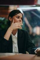 elegante mujer se sienta en un café a un mesa y fuma un cigarrillo liberando fumar desde su boca, un malo hábito, un sonrisa con dientes y un pensativo Mira foto