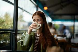 mujer sentado en un café Bebiendo café desde un jarra mirando fuera el ventana, retrato de un elegante niña con rojo pelo en el otoño foto