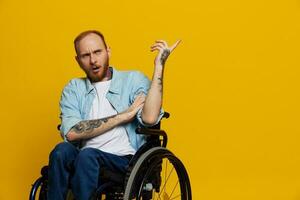 A man in a wheelchair problems with the musculoskeletal system looks at the camera shows a finger on, with tattoos on his hands sits on a yellow studio background, health concept man with disabilities photo