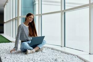 A woman works online via the Internet on her laptop and smiles looking out the large windows of a tall building, an urban style concept and working in an office photo