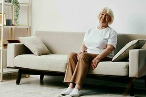 mayor mujer se sienta en sofá a hogar, brillante espacioso interior en antiguo años sonrisa, estilo de vida. abuela con gris pelo en un blanco camiseta y beige pantalones. foto