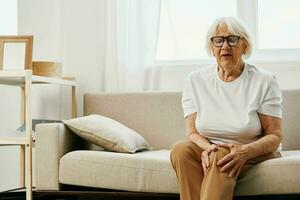 mayor mujer grave dolor en su pierna sentado en el sofá, salud problemas en antiguo edad, pobre calidad de vida. abuela con gris pelo sostiene en a su dolorido rodilla, problemas con articulaciones y ligamentos foto