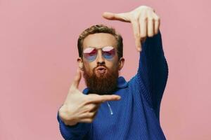 Portrait of a redheaded man wearing sunglasses smiling and dancing, listening to music on a pink background. Hipster with a beard, happiness finger pointing. photo