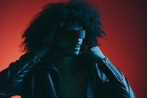 Portrait of fashion man with curly hair on red background with stylish glasses, multicultural, colored light, black leather jacket trend, modern concept. photo
