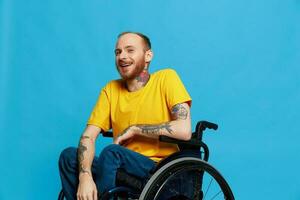 un hombre en un silla de ruedas sonrisa mira a el cámara en un camiseta con tatuajes en su brazos se sienta en un azul estudio fondo, un lleno vida, un real persona foto