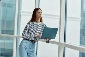 A woman standing near a large window in the city and holding a laptop in her hand looking out the window at the cityscape, a copy space photo