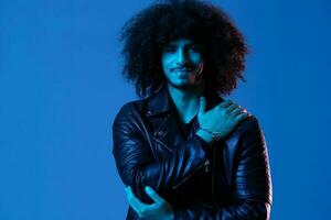 Portrait of fashion man with curly hair on blue background multinational, colored light, black leather jacket trend, modern concept. photo