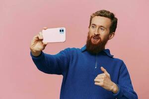 Portrait of a redheaded man with phone in hand taking selfies and photos on his phone with a smile on a pink background, blogger