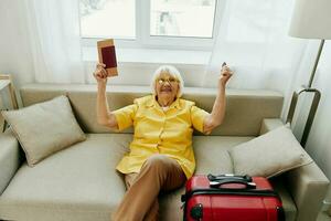 Happy senior woman with passport and travel ticket packed a red suitcase, vacation and health care. Smiling old woman joyfully sitting on the sofa before the trip raised her hands up in joy. photo