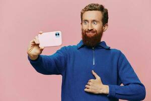 Portrait of a redheaded man with phone in hand taking selfies and photos on his phone with a smile on a pink background, blogger